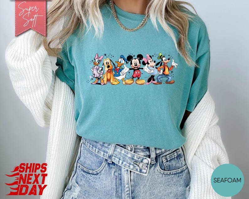 Puede incluir: Camiseta verde agua con un dise&ntilde;o de personajes de Disney: Mickey, Minnie, Donald, Daisy, Goofy y Pluto. La camiseta tiene cuello redondo y mangas cortas. Las palabras "Super Soft" est&aacute;n en una pancarta rosa. Hecha de material suave.