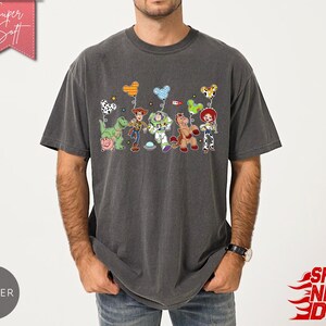 May include: A charcoal grey short-sleeve t-shirt featuring a graphic print of various animated toy characters, including a cowboy, a space ranger, a cowgirl, a dinosaur, a pig, and a horse, each holding a colourful patterned balloon shaped like mouse ears.