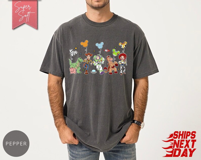 May include: A charcoal grey short-sleeve t-shirt featuring a graphic print of various animated toy characters, including a cowboy, a space ranger, a cowgirl, a dinosaur, a pig, and a horse, each holding a colourful patterned balloon shaped like mouse ears.