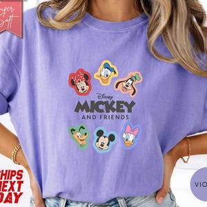 May include: A violet-colored t-shirt featuring the text "Disney MICKEY AND FRIENDS" with cartoon character graphics. The shirt has rolled-up sleeves and a relaxed fit. The design includes Mickey Mouse, Minnie Mouse, Donald Duck, Daisy Duck, and Goofy.