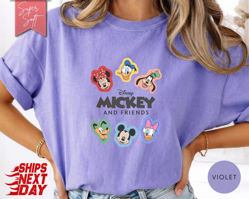 May include: A violet-colored t-shirt featuring the text "Disney MICKEY AND FRIENDS" with cartoon character graphics. The shirt has rolled-up sleeves and a relaxed fit. The design includes Mickey Mouse, Minnie Mouse, Donald Duck, Daisy Duck, and Goofy.