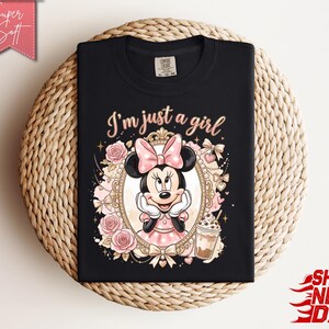 Może przedstawiać: Czarny t-shirt z grafiką Myszki Minnie w ozdobnej złotej ramce, otoczonej r&oacute;żowymi r&oacute;żami i kokardkami. Napis "I'm just a girl" jest wydrukowany jasnor&oacute;żową czcionką nad ramką, obok znajduje się ilustracja mrożonej kawy.