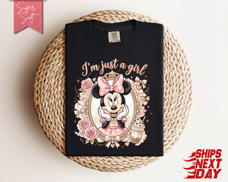Może przedstawiać: Czarny t-shirt z grafiką Myszki Minnie w ozdobnej złotej ramce, otoczonej r&oacute;żowymi r&oacute;żami i kokardkami. Napis "I'm just a girl" jest wydrukowany jasnor&oacute;żową czcionką nad ramką, obok znajduje się ilustracja mrożonej kawy.