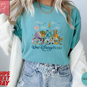 K&ouml;nnte beinhalten: T&uuml;rkisfarbenes T-Shirt mit einer farbenfrohen Grafik von Walt Disney World-Charakteren und -Wahrzeichen. Das Design zeigt Micky Maus, Donald Duck und andere Charaktere mit dem Text "Walt Disney World Since 1971".