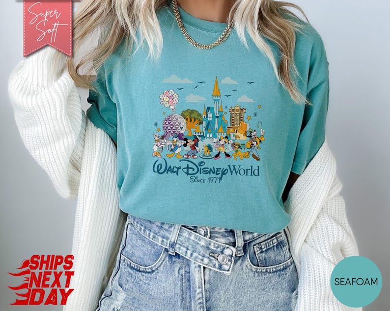K&ouml;nnte beinhalten: T&uuml;rkisfarbenes T-Shirt mit einer farbenfrohen Grafik von Walt Disney World-Charakteren und -Wahrzeichen. Das Design zeigt Micky Maus, Donald Duck und andere Charaktere mit dem Text "Walt Disney World Since 1971".
