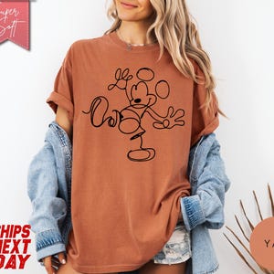 May include: A rust-colored t-shirt featuring a black line drawing of Mickey Mouse. The shirt has rolled-up sleeves and the text "Super Soft" in a pink banner. The words "Ships Next Day" are also visible.