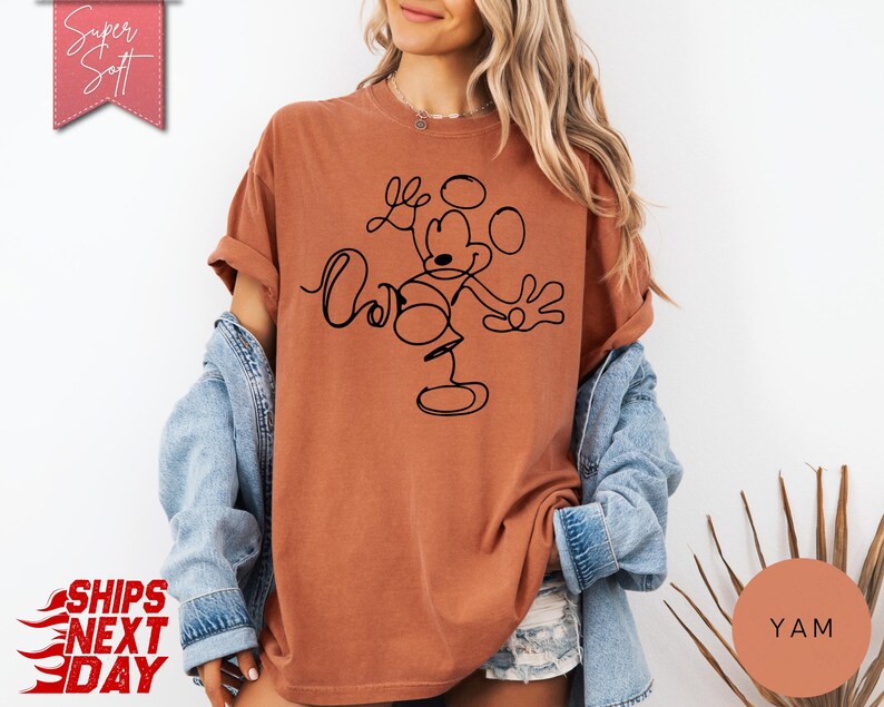 May include: A rust-colored t-shirt featuring a black line drawing of Mickey Mouse. The shirt has rolled-up sleeves and the text "Super Soft" in a pink banner. The words "Ships Next Day" are also visible.