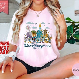 K&ouml;nnte beinhalten: Hellrosa T-Shirt mit einem farbenfrohen Aufdruck von Disney-Figuren und Wahrzeichen, darunter Cinderella's Schloss und Spaceship Earth. Der Text "Walt Disney World Since 1971" ist darunter gedruckt. Das T-Shirt hat eine lockere Passform.