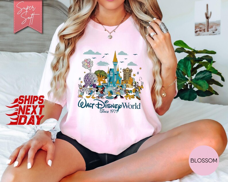 K&ouml;nnte beinhalten: Hellrosa T-Shirt mit einem farbenfrohen Aufdruck von Disney-Figuren und Wahrzeichen, darunter Cinderella's Schloss und Spaceship Earth. Der Text "Walt Disney World Since 1971" ist darunter gedruckt. Das T-Shirt hat eine lockere Passform.