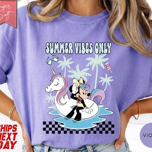 May include: A light purple t-shirt featuring a graphic print of Minnie Mouse sitting on a white unicorn pool float in blue water. The design includes palm trees, sunglasses, a popsicle, and the text SUMMER VIBES ONLY above a black and white checkerboard pattern.