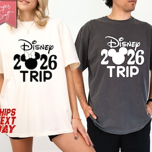 May include: Two short-sleeved t-shirts, one ivory and one pepper, featuring a Disney design. The design includes the word "Disney", the number "2026", a Mickey Mouse head, and the word "TRIP" in a bold, black font. The ivory shirt has a bow on the Mickey Mouse head.