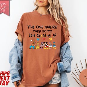 K&ouml;nnte beinhalten: Ein rostfarbenes T-Shirt mit dem Text "The One Where They Go To Disney" in Schwarz, wobei das Wort "Disney" in einer verspielten Schriftart gehalten ist. Darunter befinden sich Zeichentrickfiguren wie Mickey und Minnie Maus. Das Shirt hat hochgekrempelte &Auml;rmel.