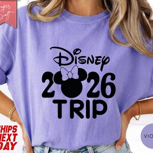 May include: A violet-colored t-shirt with the text "Disney 2026 TRIP" in black. The "0" in "2026" is replaced with a silhouette of Minnie Mouse's head and bow. The shirt has rolled-up sleeves and a "Super Soft" label.