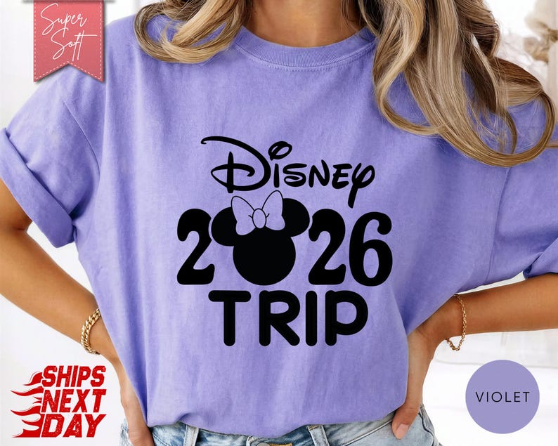 May include: A violet-colored t-shirt with the text "Disney 2026 TRIP" in black. The "0" in "2026" is replaced with a silhouette of Minnie Mouse's head and bow. The shirt has rolled-up sleeves and a "Super Soft" label.