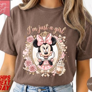 Może przedstawiać: Brązowy t-shirt z grafiką Myszki Minnie w ozdobnej złotej owalnej ramce. Nad ramką nadrukowano napis "I'm just a girl". Wz&oacute;r zawiera r&oacute;żowe r&oacute;że, kokardki oraz kubek ze słomką. R&oacute;żowa metka zawiera napis "Super Soft".