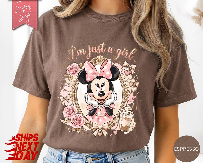 Może przedstawiać: Brązowy t-shirt z grafiką Myszki Minnie w ozdobnej złotej owalnej ramce. Nad ramką nadrukowano napis "I'm just a girl". Wz&oacute;r zawiera r&oacute;żowe r&oacute;że, kokardki oraz kubek ze słomką. R&oacute;żowa metka zawiera napis "Super Soft".