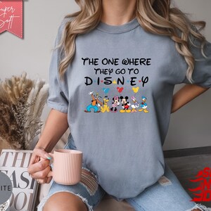 K&ouml;nnte beinhalten: Ein hellblaues T-Shirt mit dem Text "The One Where They Go To Disney" und Disney-Figuren. Das Shirt hat einen Rundhalsausschnitt und kurze &Auml;rmel. Das Design umfasst Micky Maus, Minnie Maus, Donald Duck und Goofy.