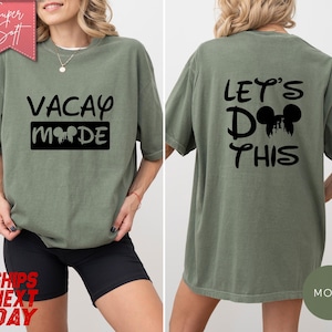 May include: A moss green t-shirt with the words "VACAY MODE" printed on the front in black. The back of the shirt says "LET'S DO THIS" with a Mickey Mouse silhouette. The shirt is a relaxed fit.