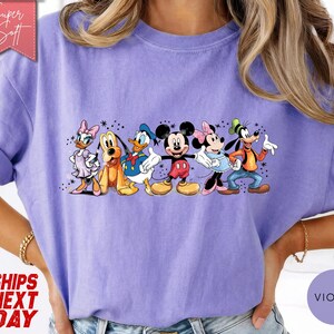 Puede incluir: Una camiseta de color violeta con un gr&aacute;fico de personajes de Disney, incluyendo Mickey Mouse, Minnie Mouse, Donald Duck y Goofy. El dise&ntilde;o est&aacute; centrado en la camiseta. La camiseta tiene mangas enrolladas.