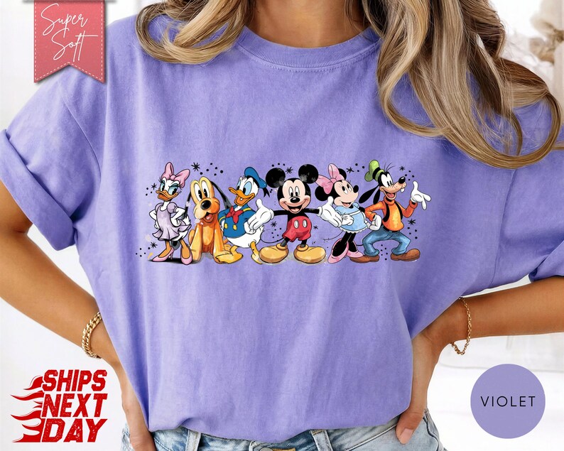 Puede incluir: Una camiseta de color violeta con un gr&aacute;fico de personajes de Disney, incluyendo Mickey Mouse, Minnie Mouse, Donald Duck y Goofy. El dise&ntilde;o est&aacute; centrado en la camiseta. La camiseta tiene mangas enrolladas.