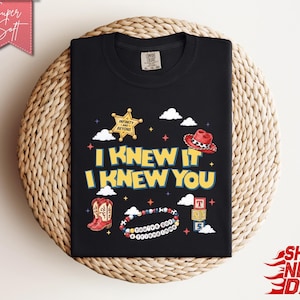 May include: A black t-shirt featuring a graphic print with the text I KNEW IT I KNEW YOU in yellow letters. The design includes a gold sheriff star, a red cowboy hat, red cowboy boots, a beaded bracelet with the text YOU'VE GOT A FRIEND IN ME.
