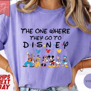K&ouml;nnte beinhalten: Violettes T-Shirt mit dem schwarzen Schriftzug "THE ONE WHERE THEY GO TO DISNEY". Unter dem Text sind Zeichentrickfiguren mit Luftballons abgebildet. Das T-Shirt hat kurze &Auml;rmel und eine lockere Passform.