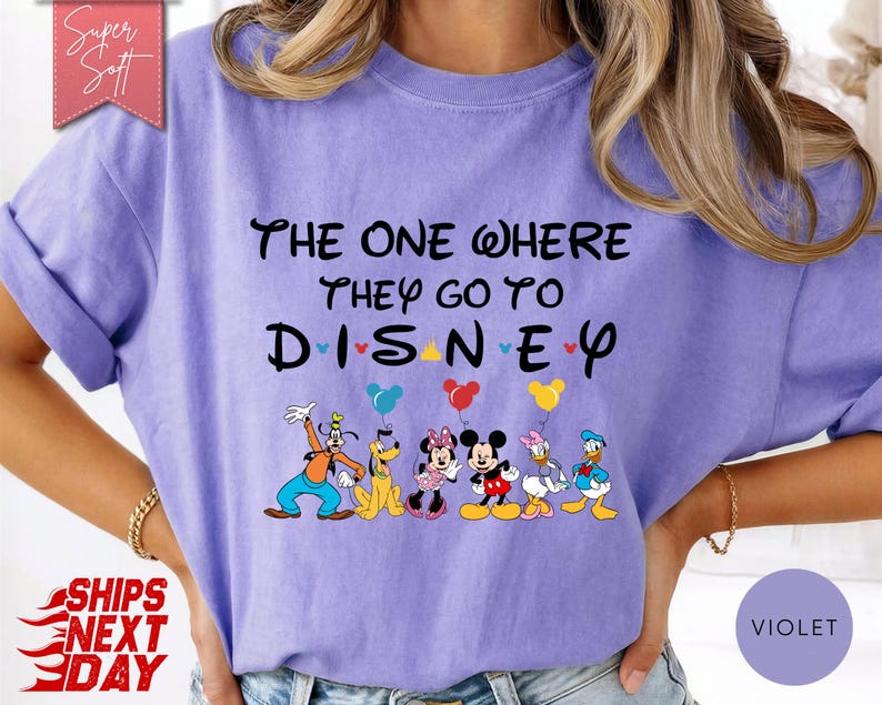 K&ouml;nnte beinhalten: Violettes T-Shirt mit dem schwarzen Schriftzug "THE ONE WHERE THEY GO TO DISNEY". Unter dem Text sind Zeichentrickfiguren mit Luftballons abgebildet. Das T-Shirt hat kurze &Auml;rmel und eine lockere Passform.