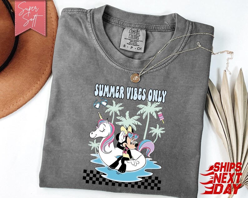 May include: A charcoal gray short-sleeve t-shirt featuring a graphic print of a cartoon mouse and a unicorn on a pool float. The text reads SUMMER VIBES ONLY. The design includes palm trees, heart-shaped sunglasses, and a popsicle, with a black and white checkered pattern at the bottom.