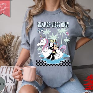 May include: A slate blue t-shirt featuring a graphic print with the text "SUMMER VIBES ONLY". The design includes a cartoon mouse character sitting on a white unicorn pool float in blue water, surrounded by palm trees, sunglasses, and a popsicle.