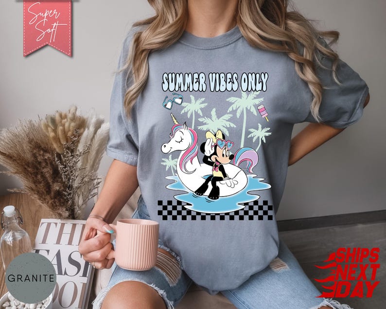 May include: A slate blue t-shirt featuring a graphic print with the text "SUMMER VIBES ONLY". The design includes a cartoon mouse character sitting on a white unicorn pool float in blue water, surrounded by palm trees, sunglasses, and a popsicle.