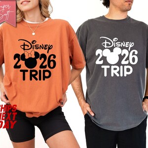 May include: Two short-sleeved t-shirts, one orange and one gray, each with a Disney design. The design includes the Disney logo, the number 2026, and the word "TRIP". The orange shirt has a Minnie Mouse bow.