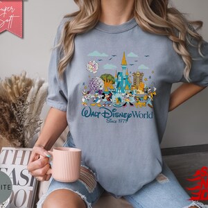 K&ouml;nnte beinhalten: Hellblaues T-Shirt mit farbenfroher Grafik von Walt Disney World, mit Micky Maus und Freunden. Das Shirt hat den Text "Walt Disney World Since 1971". Das Shirt ist aus weichem Material.