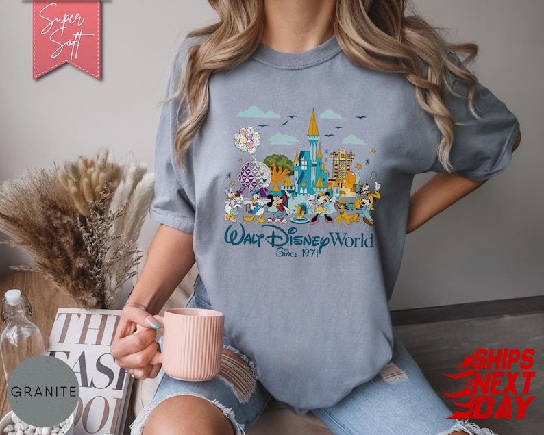 K&ouml;nnte beinhalten: Hellblaues T-Shirt mit farbenfroher Grafik von Walt Disney World, mit Micky Maus und Freunden. Das Shirt hat den Text "Walt Disney World Since 1971". Das Shirt ist aus weichem Material.