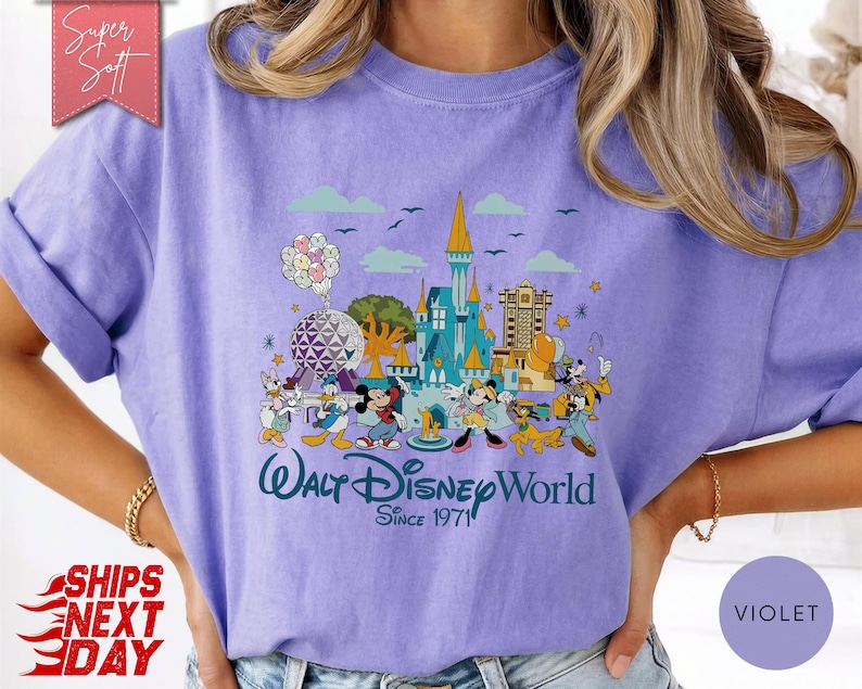 K&ouml;nnte beinhalten: Ein violettes T-Shirt mit einer farbenfrohen Grafik von Disney-Figuren und Wahrzeichen. Das Design umfasst Micky Maus, Donald Duck und ein Schloss mit dem Text "Walt Disney World Since 1971". Das Shirt hat kurze &Auml;rmel und eine lockere Passform.