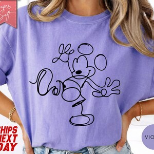 May include: A violet-colored t-shirt featuring a black line drawing of Mickey Mouse. The design is a continuous line, creating a minimalist style. The shirt has rolled-up sleeves and a relaxed fit. The text "Super Soft" is in a pink banner.