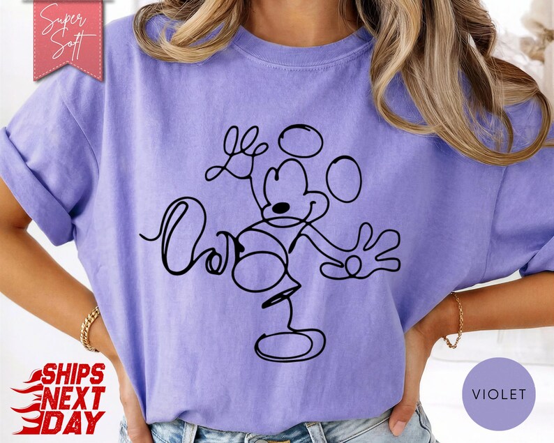 May include: A violet-colored t-shirt featuring a black line drawing of Mickey Mouse. The design is a continuous line, creating a minimalist style. The shirt has rolled-up sleeves and a relaxed fit. The text "Super Soft" is in a pink banner.