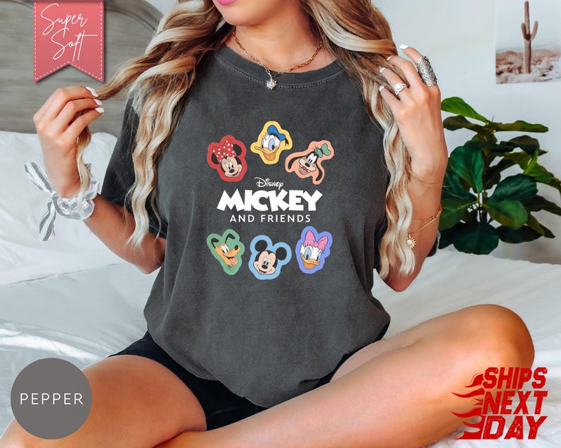 May include: Dark gray t-shirt featuring the text "Mickey and Friends" with colorful character graphics. The shirt has a crew neck and short sleeves. The image also includes the text "Super Soft" and "Ships Next Day".