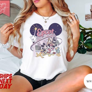 May include: White t-shirt featuring a colorful graphic of Disney characters, including Mickey Mouse, Donald Duck, and Goofy, with the word "Epcot" in a stylized font. The shirt has a round neckline and short sleeves.