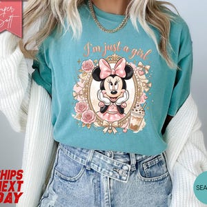 Może przedstawiać: Morskozielony t-shirt z grafiką Myszki Minnie w ozdobnej złotej ramce z r&oacute;żowymi r&oacute;żami i kokardkami. Tekst nad postacią brzmi I'm just a girl napisaną kursywą. Po prawej stronie znajduje się mały nap&oacute;j kawowy z lodem.