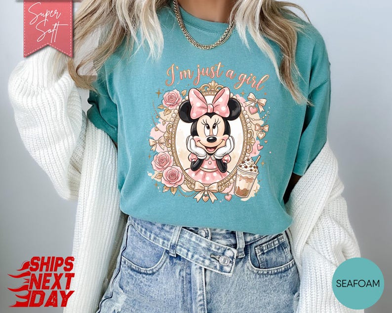 Może przedstawiać: Morskozielony t-shirt z grafiką Myszki Minnie w ozdobnej złotej ramce z r&oacute;żowymi r&oacute;żami i kokardkami. Tekst nad postacią brzmi I'm just a girl napisaną kursywą. Po prawej stronie znajduje się mały nap&oacute;j kawowy z lodem.