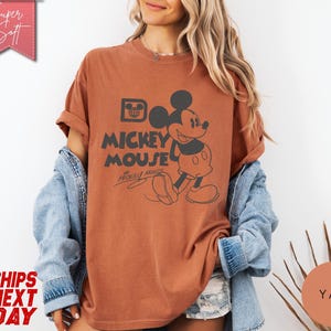 May include: A rust-colored t-shirt featuring a graphic of Mickey Mouse with the text "Mickey Mouse". The design includes a vintage-style illustration of Mickey Mouse and his signature. The shirt has rolled-up sleeves and a relaxed fit.