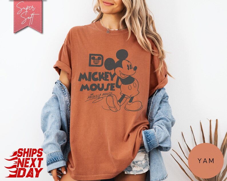 May include: A rust-colored t-shirt featuring a graphic of Mickey Mouse with the text "Mickey Mouse". The design includes a vintage-style illustration of Mickey Mouse and his signature. The shirt has rolled-up sleeves and a relaxed fit.