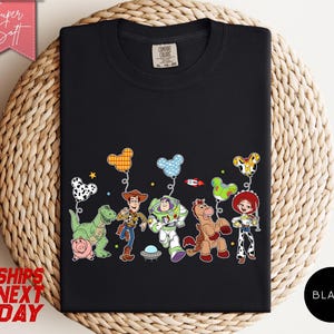 May include: A black t-shirt featuring a graphic print of animated characters including a cowboy, a space ranger, a cowgirl, a horse, a dinosaur, and a pig. Above the characters are several mouse-ear shaped balloons in various patterns and colours.
