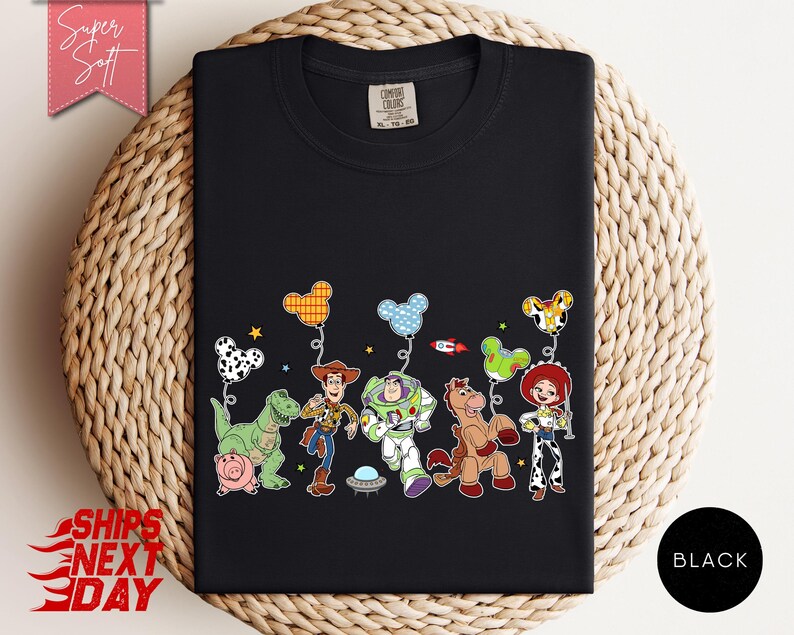 May include: A black t-shirt featuring a graphic print of animated characters including a cowboy, a space ranger, a cowgirl, a horse, a dinosaur, and a pig. Above the characters are several mouse-ear shaped balloons in various patterns and colours.
