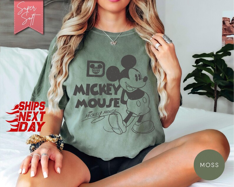 May include: An olive green t-shirt featuring a graphic of Mickey Mouse with the words "Mickey Mouse" in a vintage style. The shirt has a relaxed fit and is made of a soft material. The text "Super Soft" and "Ships Next Day" are also visible.