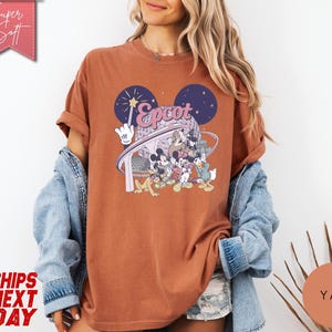 May include: A rust-colored t-shirt featuring a graphic of Mickey Mouse and friends at Epcot. The design includes the word "Epcot" in a stylized font, with a backdrop of fireworks and a futuristic structure. The shirt has rolled-up sleeves.