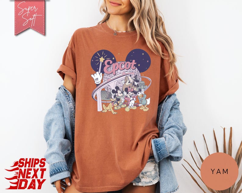 May include: A rust-colored t-shirt featuring a graphic of Mickey Mouse and friends at Epcot. The design includes the word "Epcot" in a stylized font, with a backdrop of fireworks and a futuristic structure. The shirt has rolled-up sleeves.