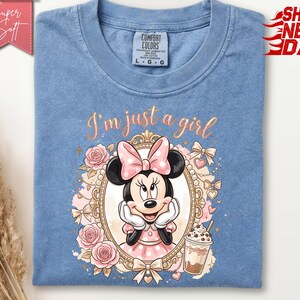 Może przedstawiać: Niebieska koszulka z okrągłym dekoltem z grafiką Myszki Minnie w ozdobnej złotej owalnej ramce. Wz&oacute;r zawiera r&oacute;żowe r&oacute;że, kokardki, kubek kawy oraz napis I'm just a girl kursywą. Koszulka posiada metkę marki Comfort Colors.