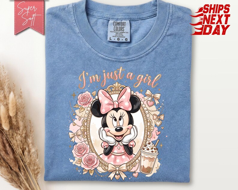 Może przedstawiać: Niebieska koszulka z okrągłym dekoltem z grafiką Myszki Minnie w ozdobnej złotej owalnej ramce. Wz&oacute;r zawiera r&oacute;żowe r&oacute;że, kokardki, kubek kawy oraz napis I'm just a girl kursywą. Koszulka posiada metkę marki Comfort Colors.