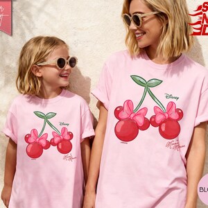 May include: Two light pink t-shirts featuring a cherry design with Minnie Mouse bows and the Disney logo. The shirts are worn by a woman and a child, both wearing sunglasses. The shirts are labeled "Super Soft" and "Blossom".
